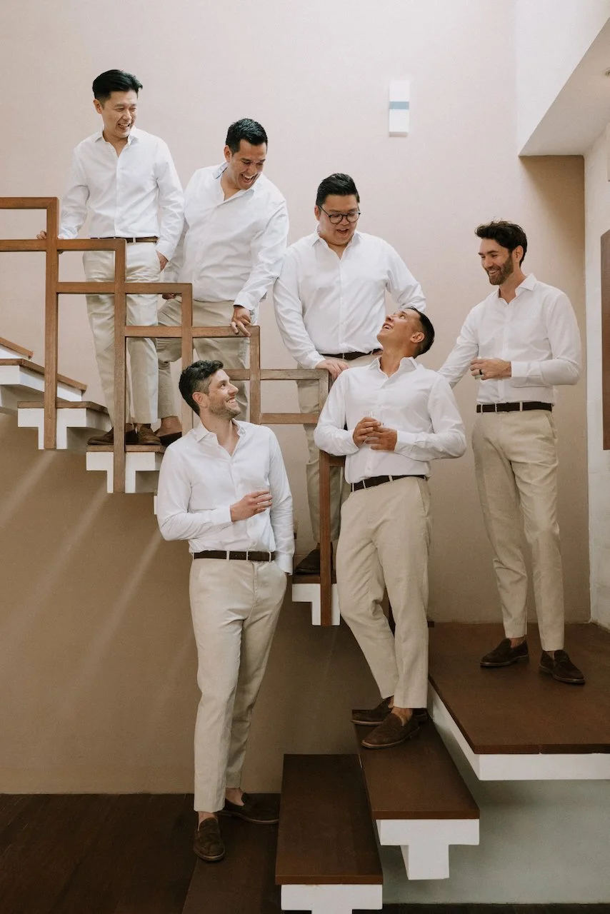 Groom and groomsmen in relaxed linen suits sharing a laugh on a modern wooden staircase.