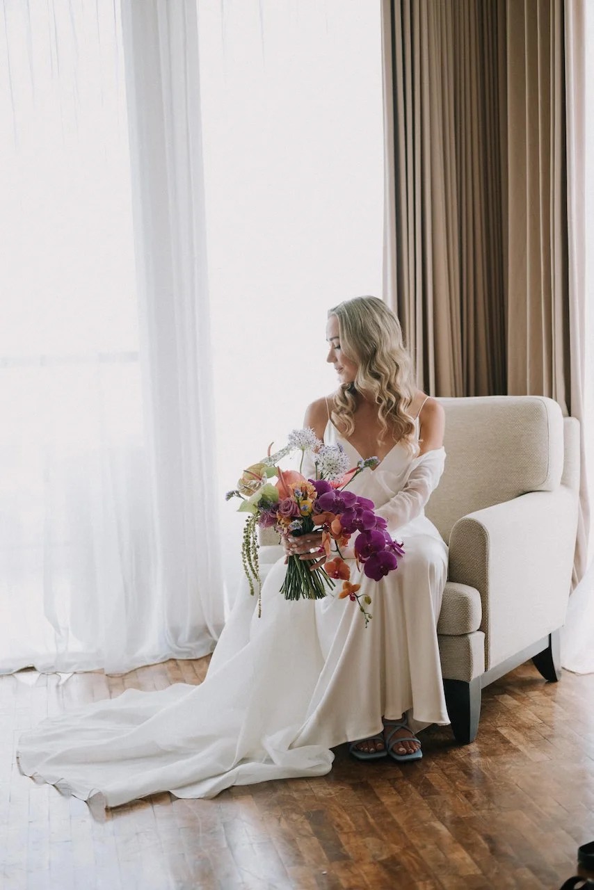 Elegant bride in a silk slip dress holding a vibrant tropical orchid bouquet by a sunlit window.