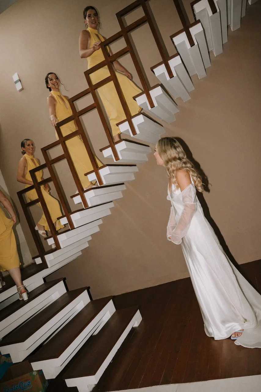 Joyful bridesmaids in yellow dresses reacting to the bride in her wedding gown at the bottom of the stairs.