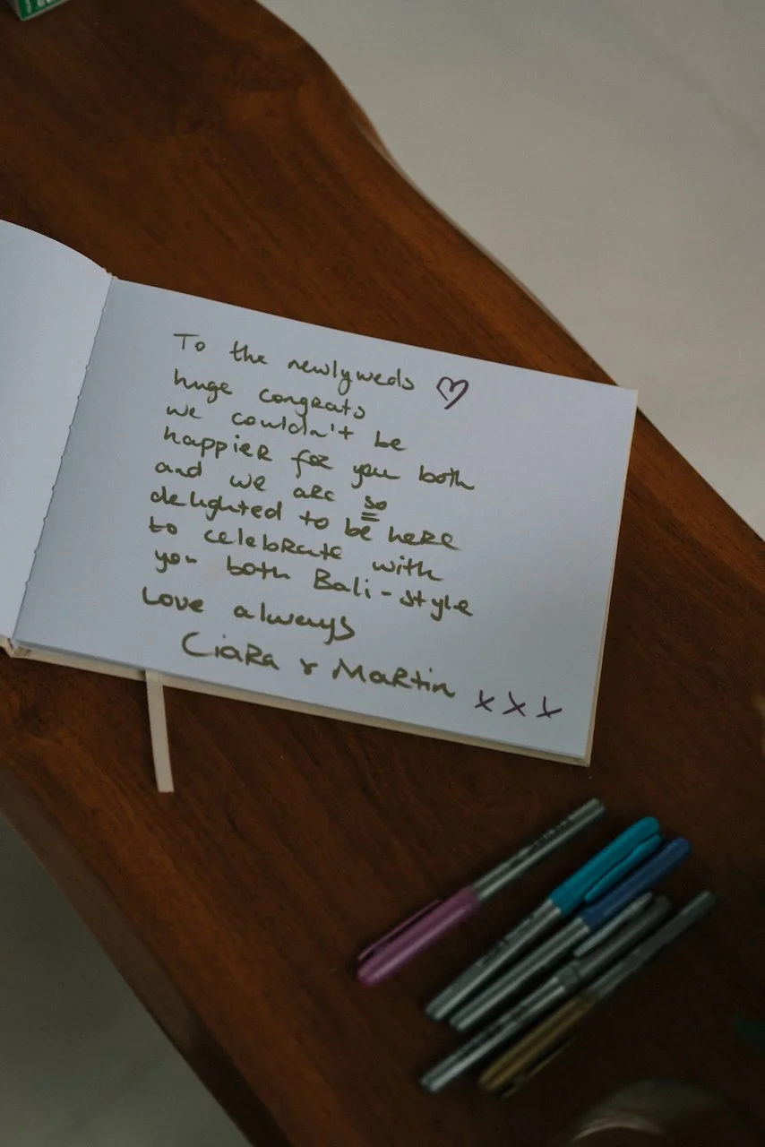 Handwritten destination wedding guest book message and colorful pens on a warm wooden table.