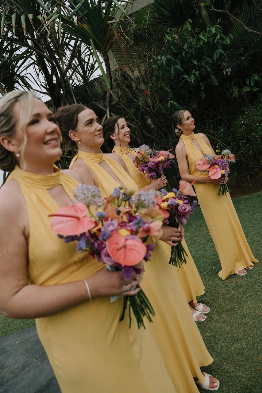 Smiling bridesmaids in flowing yellow dresses holding vibrant anthurium and orchid bouquets.