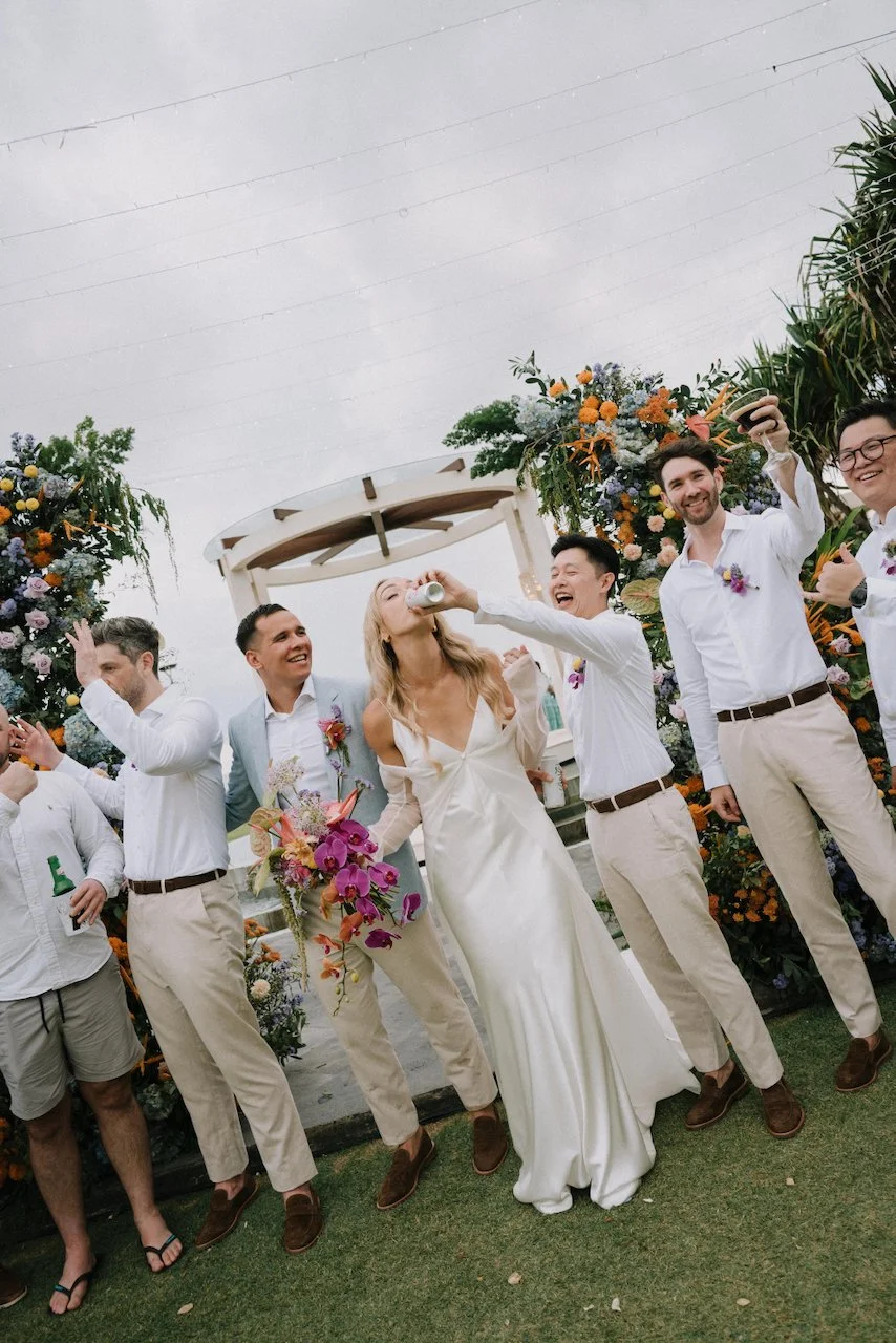 Bridal party celebrating with drinks beneath a vibrant floral arch at a Seminyak destination wedding.