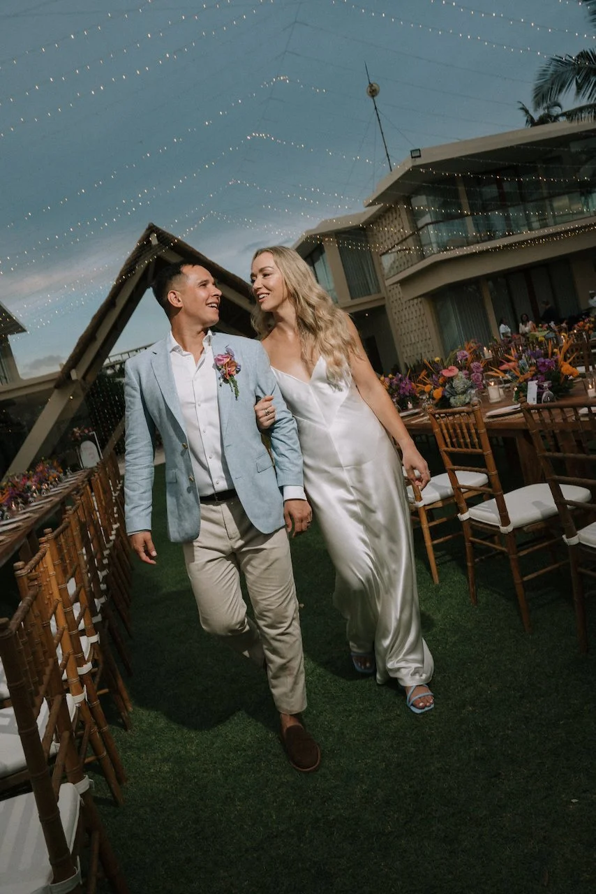 Newlyweds walking arm-in-arm past wooden reception tables under fairy lights at Villa Phalosa Bali.