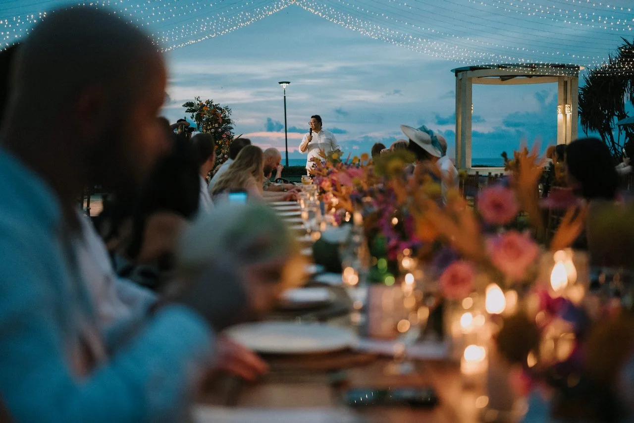 Wedding guest giving a twilight reception speech at a long floral-lined table overlooking the ocean.