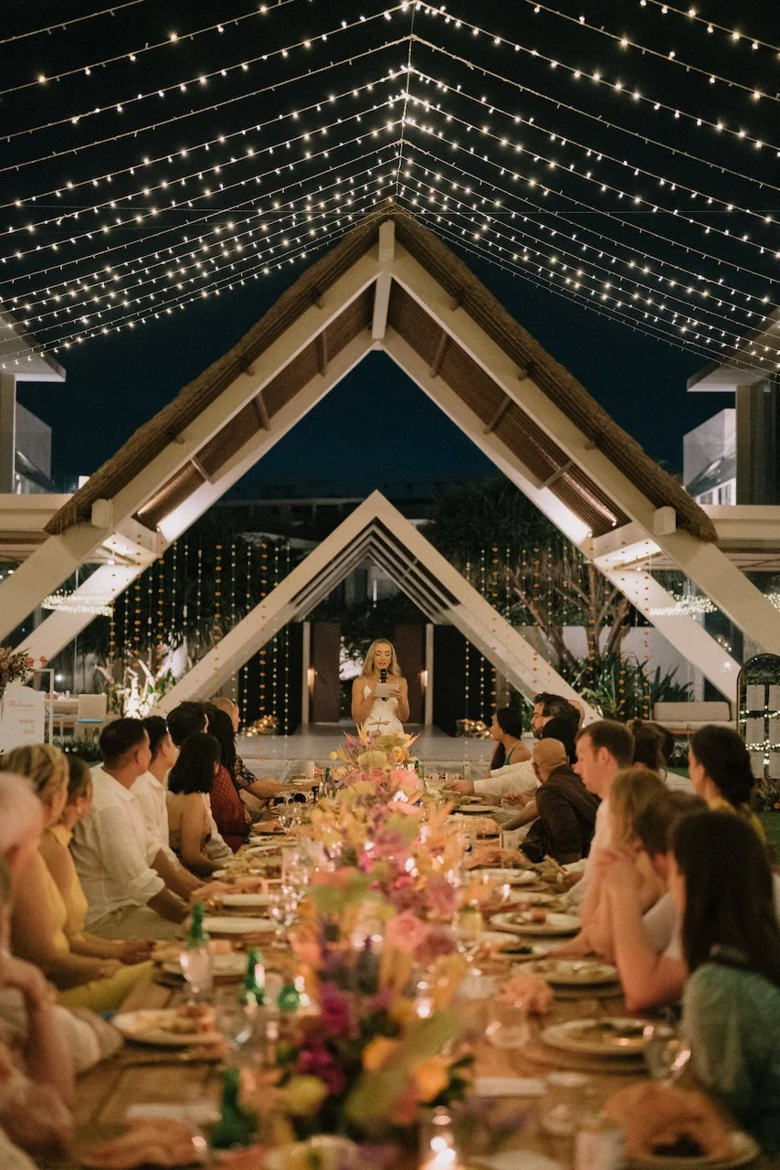 Bride delivering a touching speech under a modern A-frame structure glowing with fairy lights in Bali.