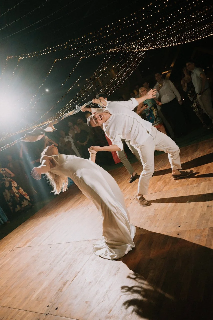 Dramatic first dance dip on an outdoor wooden floor under a canopy of romantic string lights.