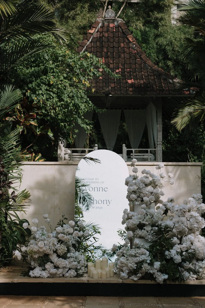 Elegant white arched wedding welcome sign surrounded by lush white florals and candles.