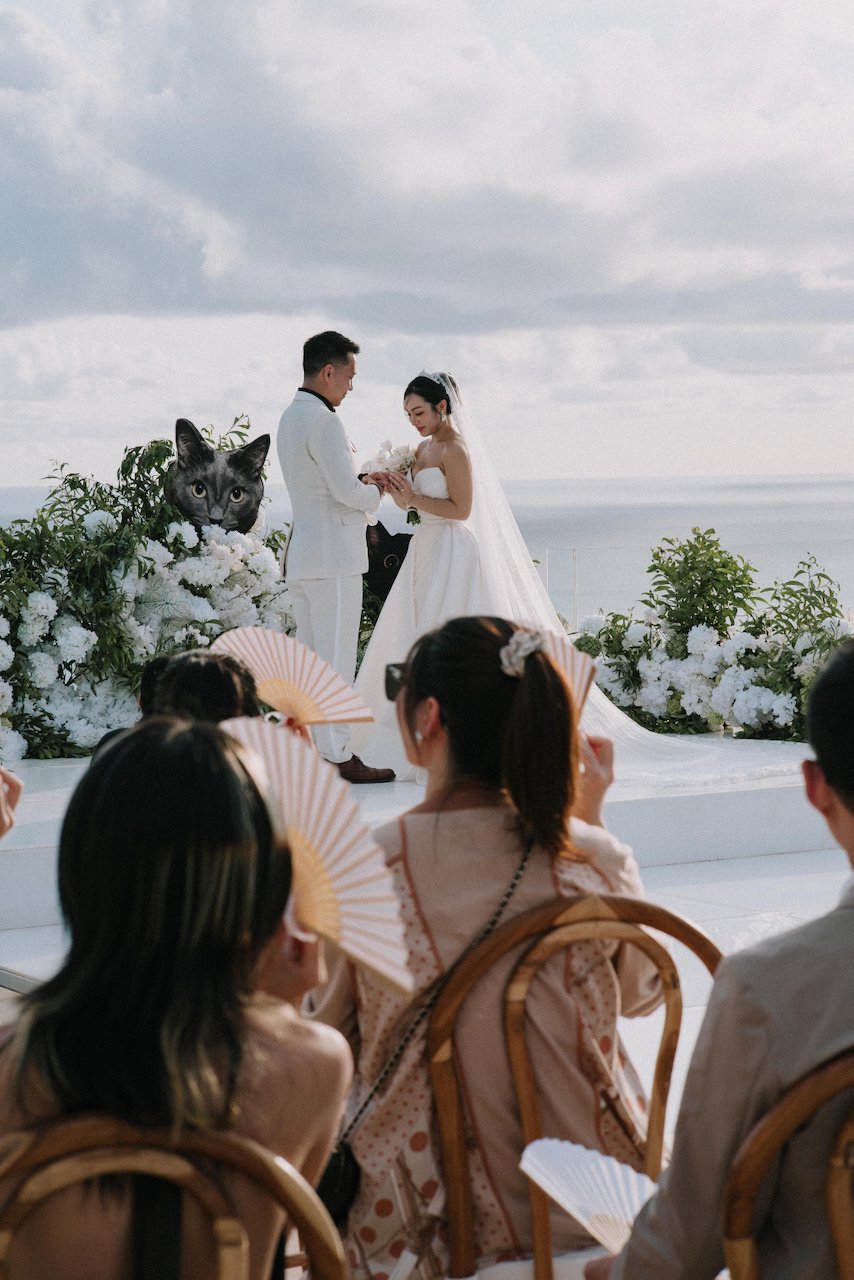 Intimate ring exchange during a clifftop wedding ceremony in Uluwatu as guests hold paper fans.