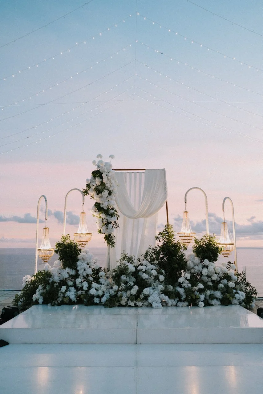 Sophisticated outdoor wedding reception stage at dusk featuring white florals, draped fabric, and crystal chandeliers.