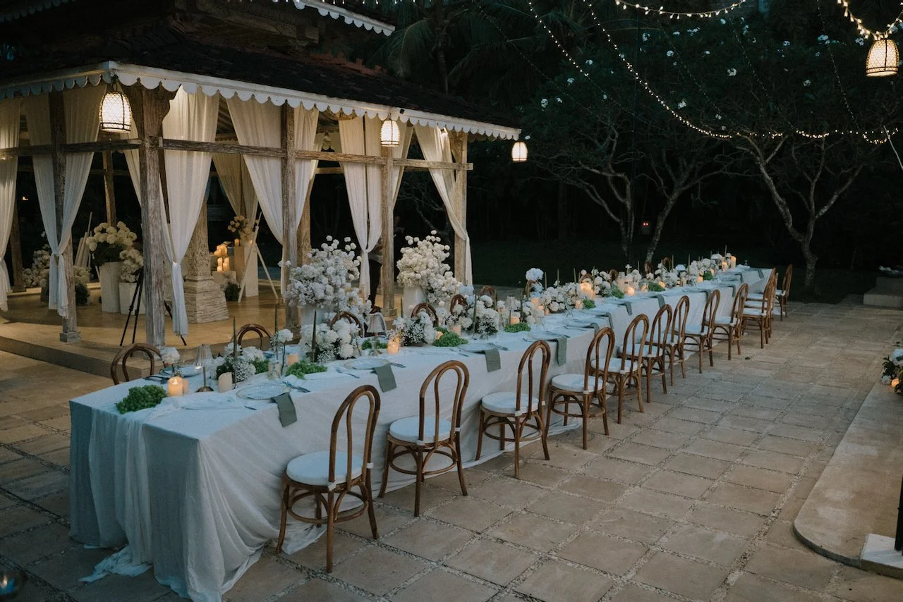 Long banquet wedding reception table decorated with lush white floral centerpieces and soft candlelight at dusk.