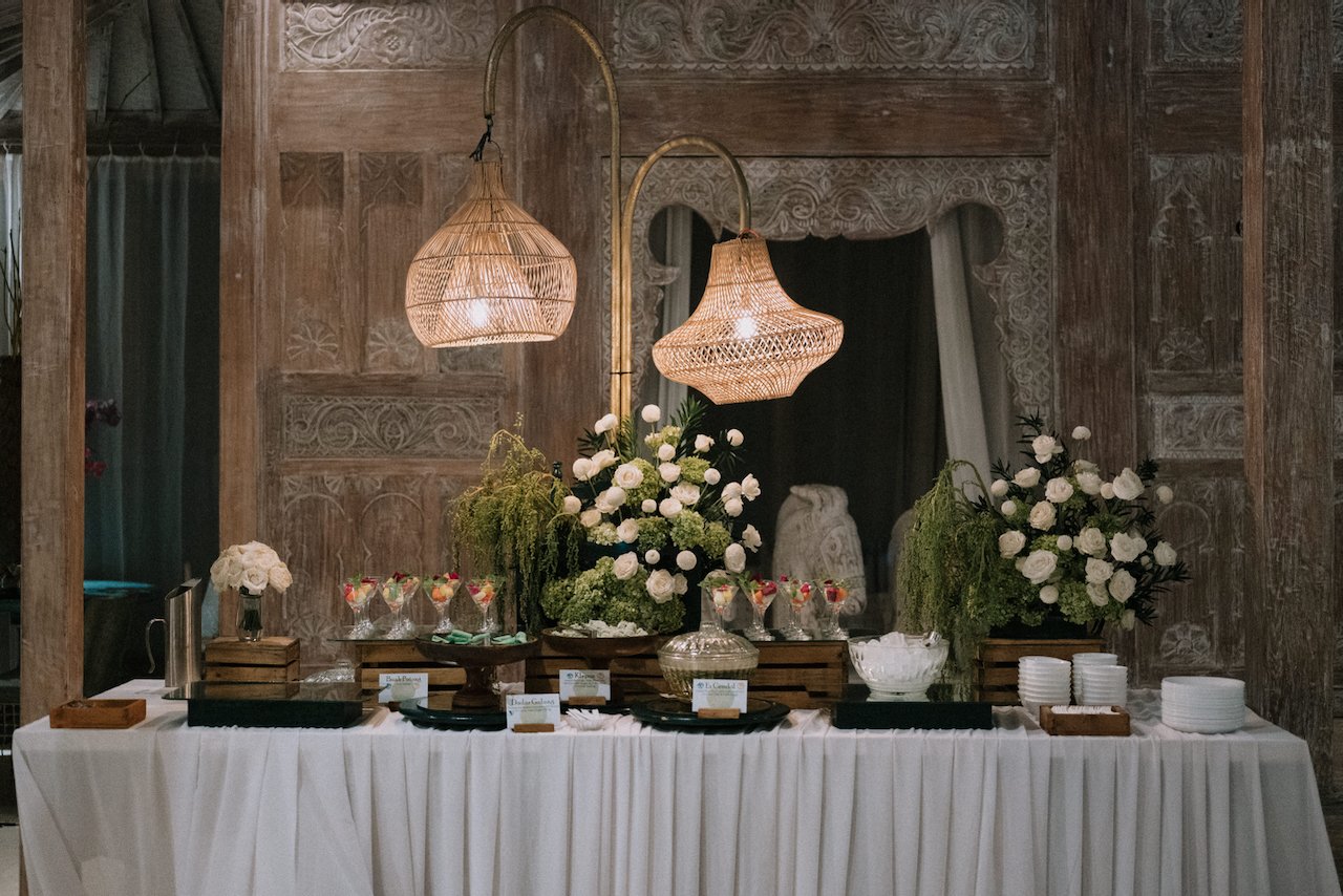 Balinese wedding dessert station featuring tropical treats, white florals, and woven rattan pendant lamps.