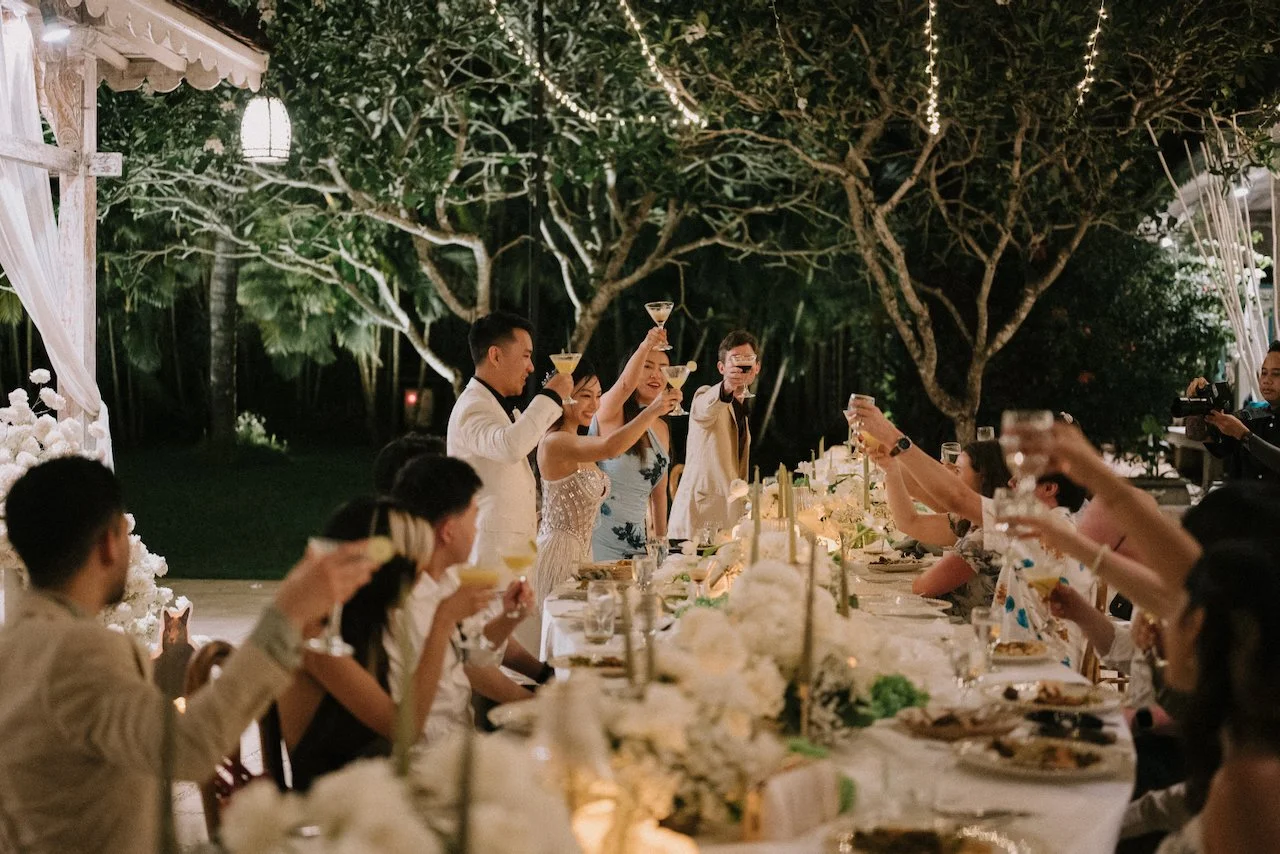 Wedding guests raising their cocktail glasses for a celebratory toast at a long outdoor dining table under the stars.