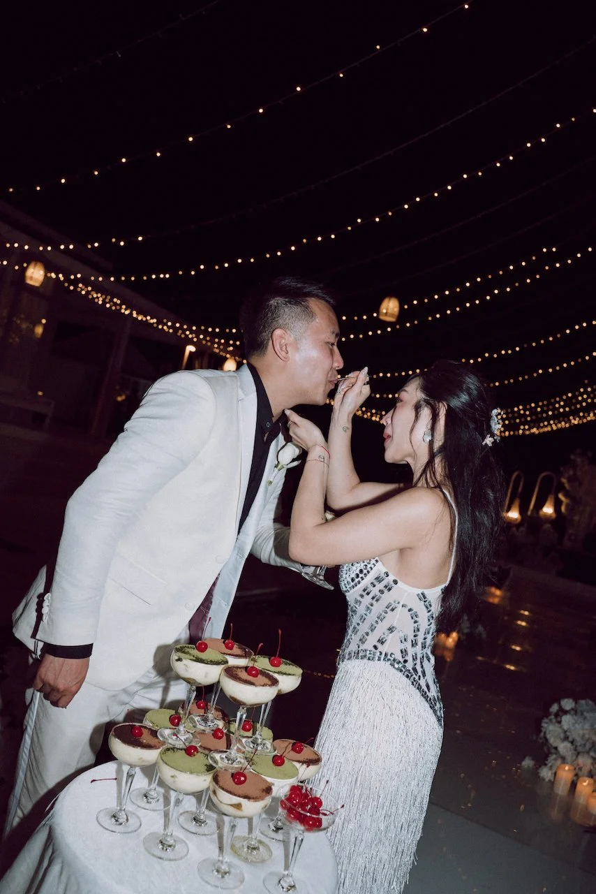 Bride in a beaded reception gown playfully feeding the groom a sweet treat from their alternative wedding dessert tower.