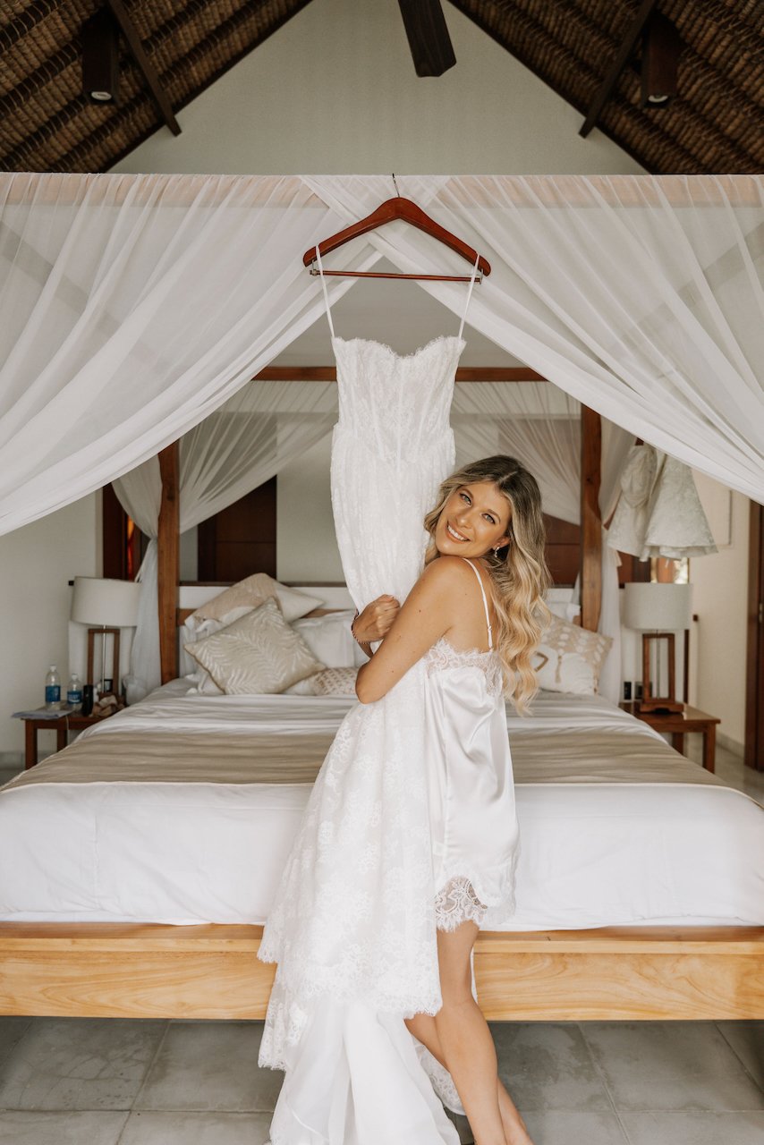 Bride smiling in a tropical villa bedroom holding up her silver lace wedding gown.