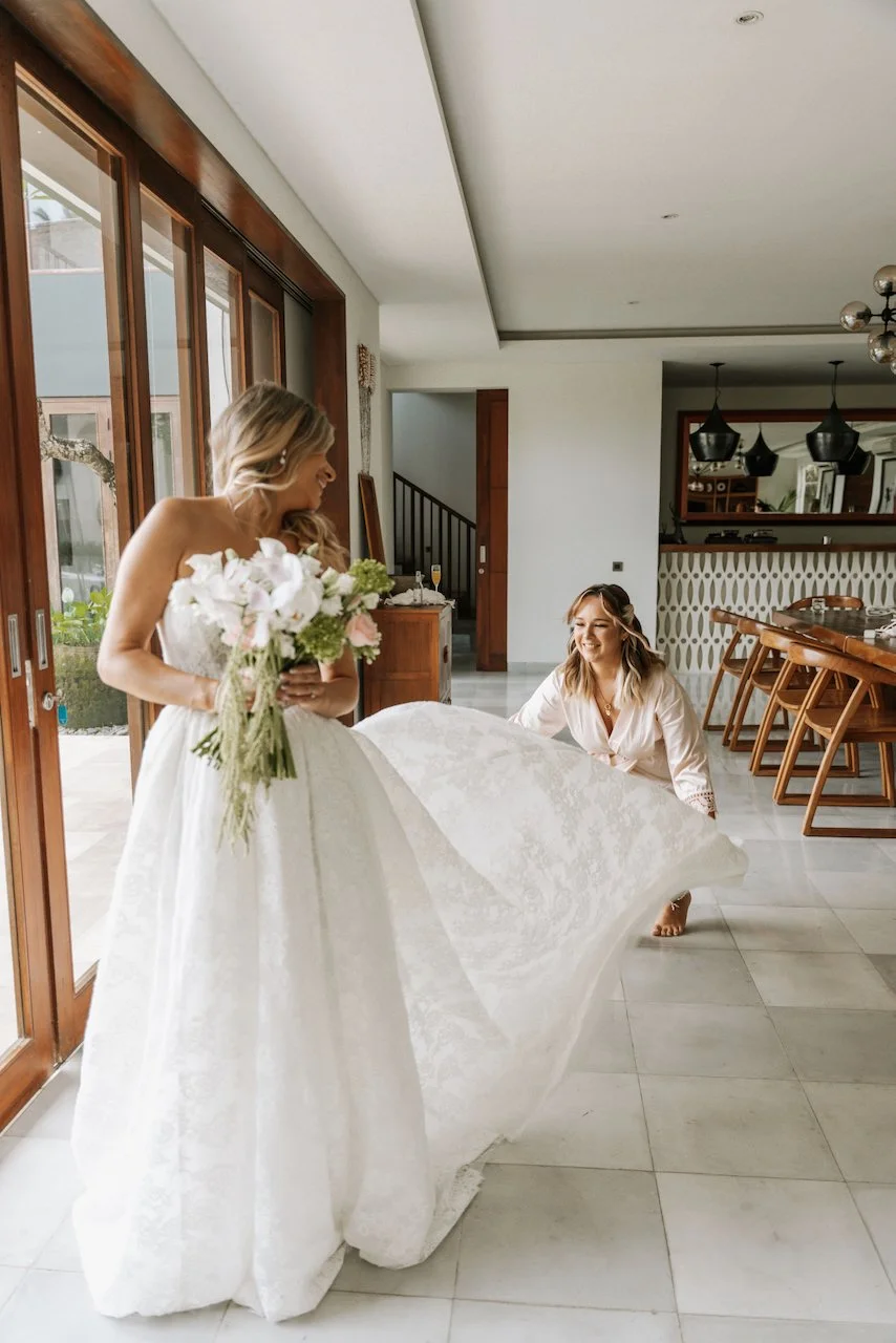 Bridesmaid helping to arrange the flowing train of a strapless wedding dress indoors.
