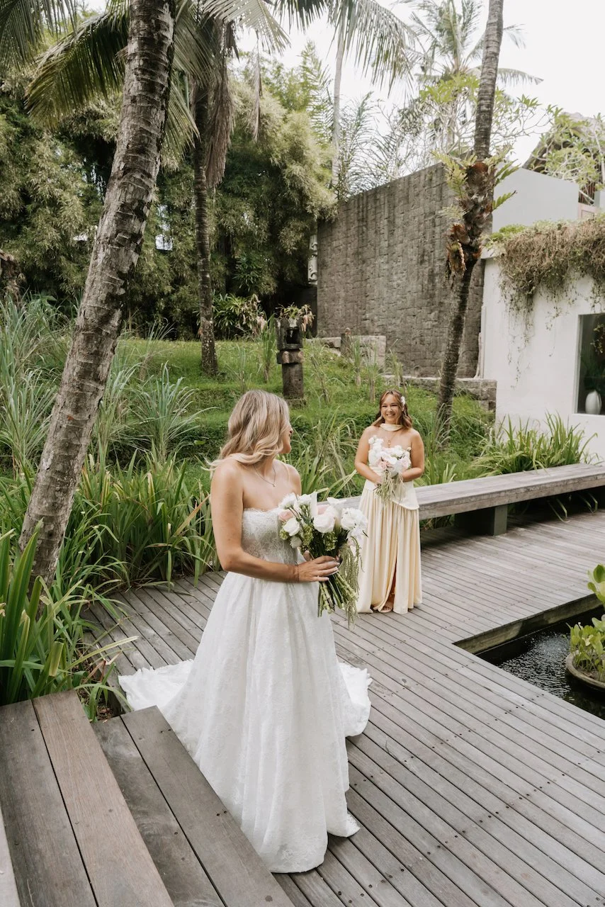 Bride and bridesmaid standing on a wooden walkway surrounded by lush tropical garden greenery.
