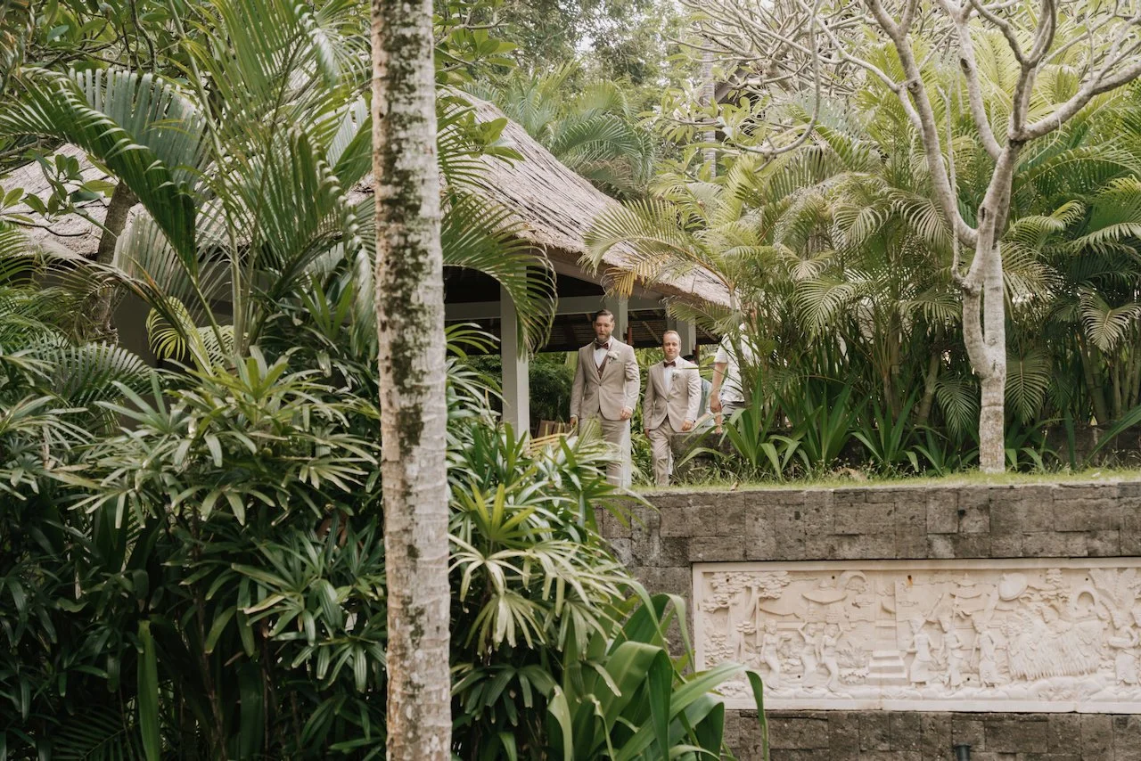 Groom and groomsman in beige suits walking through a tropical Bali garden setting.