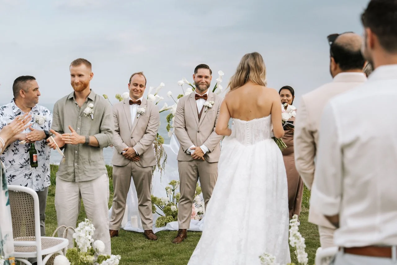 Groom smiling warmly at his bride as she walks down the aisle at an outdoor cliffside Bali wedding.