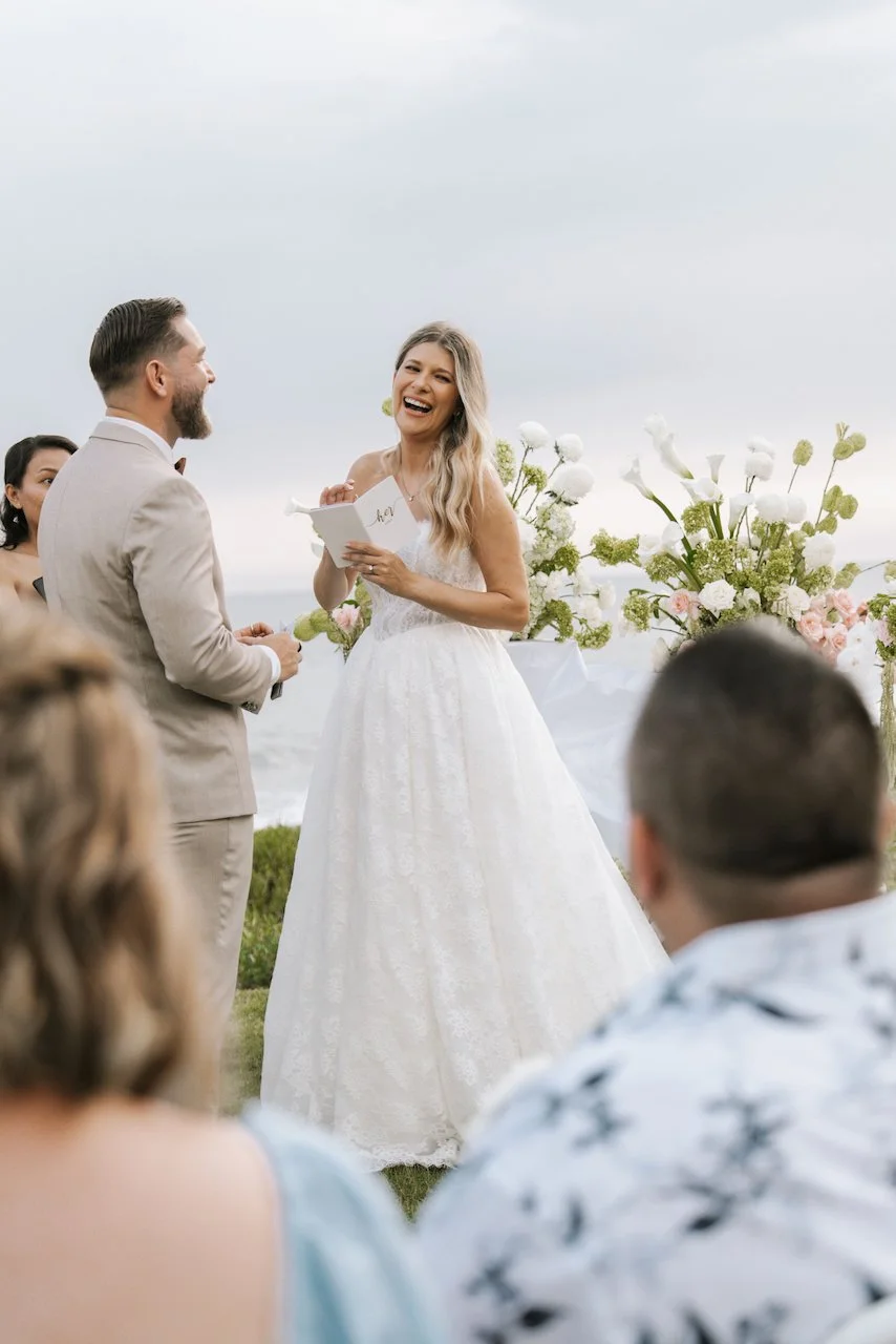 Joyful bride laughing while reading her personalized vows at a scenic oceanfront ceremony.