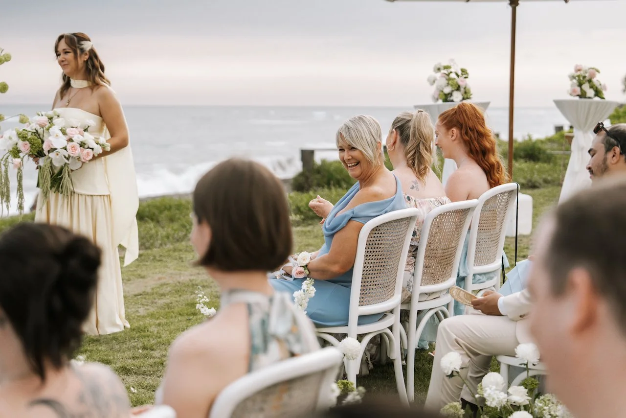 Wedding guests seated in white rattan chairs smiling warmly during a tropical ocean-view ceremony.