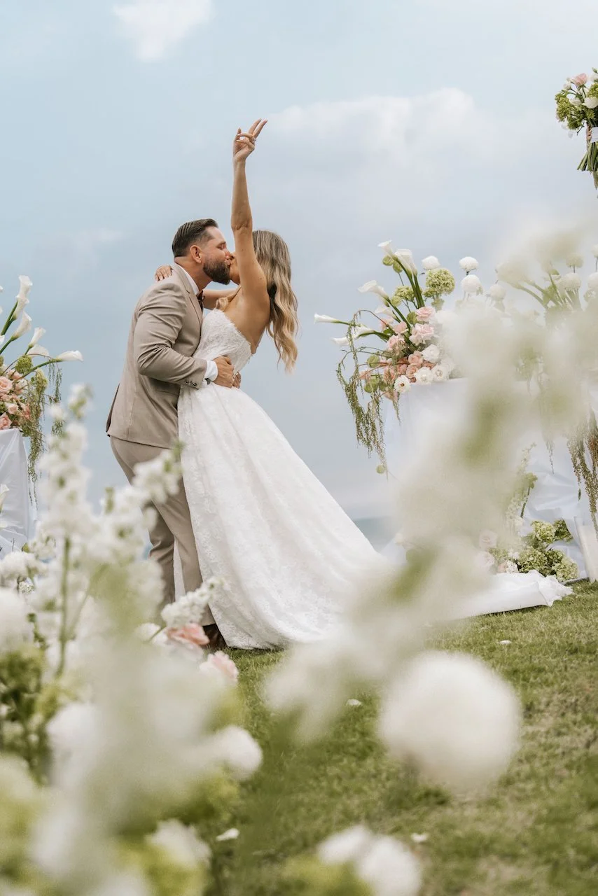Newlyweds sharing a celebratory first kiss framed by out-of-focus white wedding flowers.