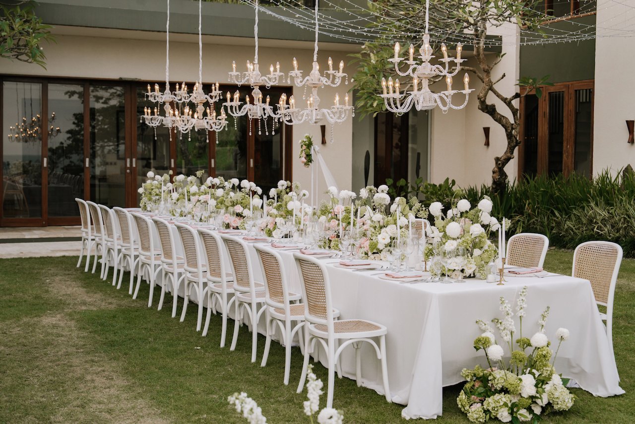 Elegant outdoor wedding reception featuring a long banquet table under hanging crystal chandeliers.