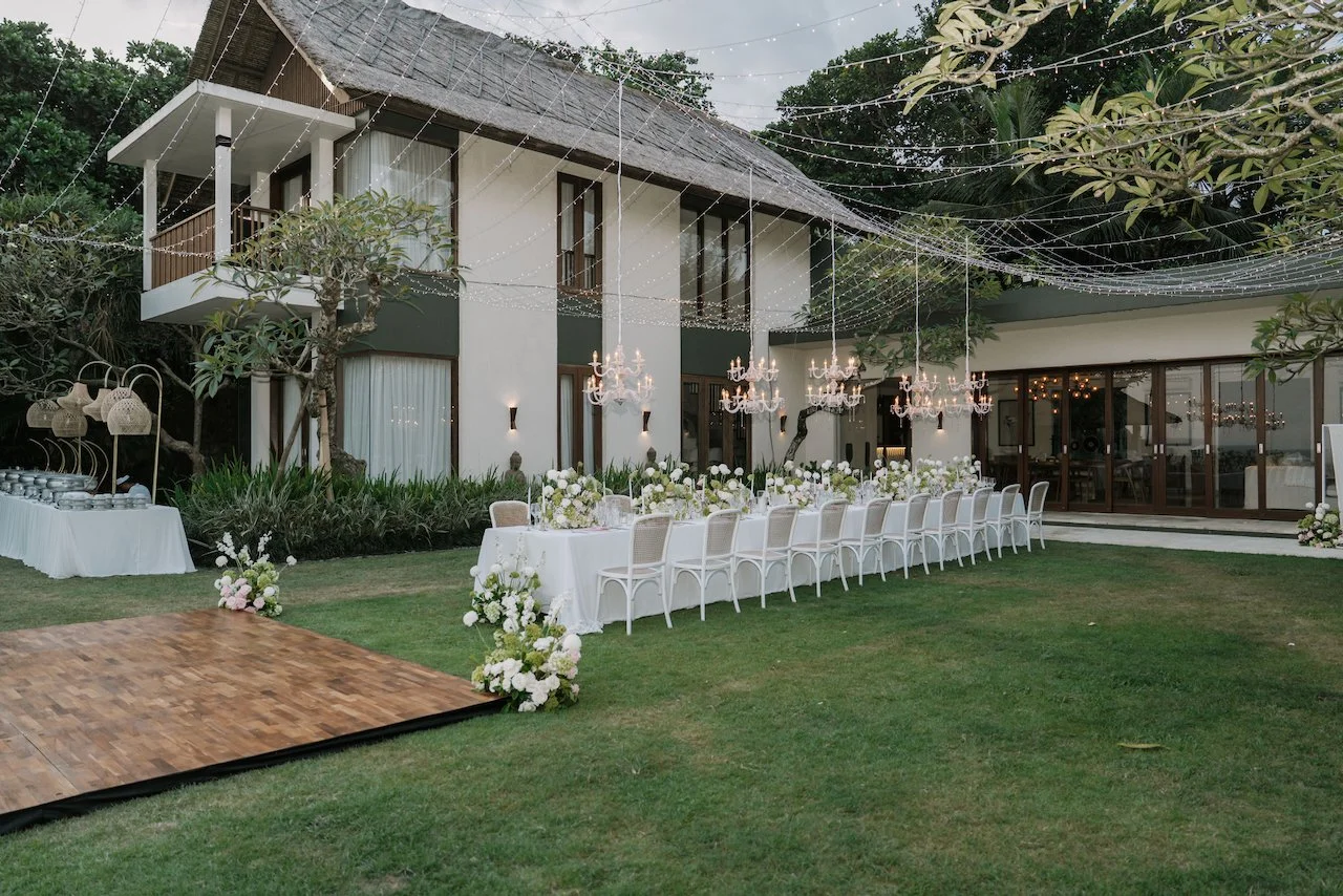 Outdoor villa wedding reception setup showcasing a long dining table, wooden dance floor, and romantic string lights.
