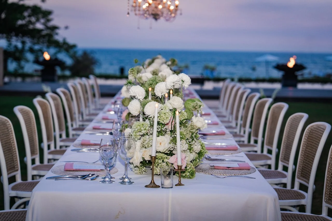 Elegant oceanfront wedding reception table at sunset featuring tall taper candles and white floral centerpieces.