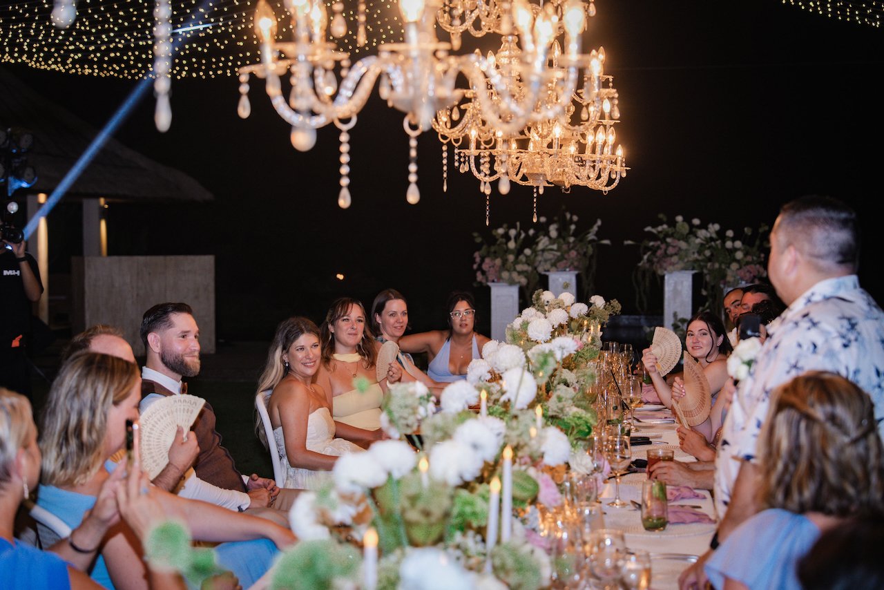 Wedding guests and newlyweds laughing during dinner speeches under outdoor crystal chandeliers at night.