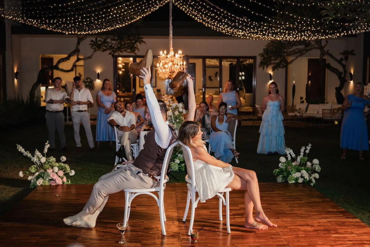 Bride and groom playing the wedding shoe game back-to-back on a wooden dance floor illuminated by fairy lights.