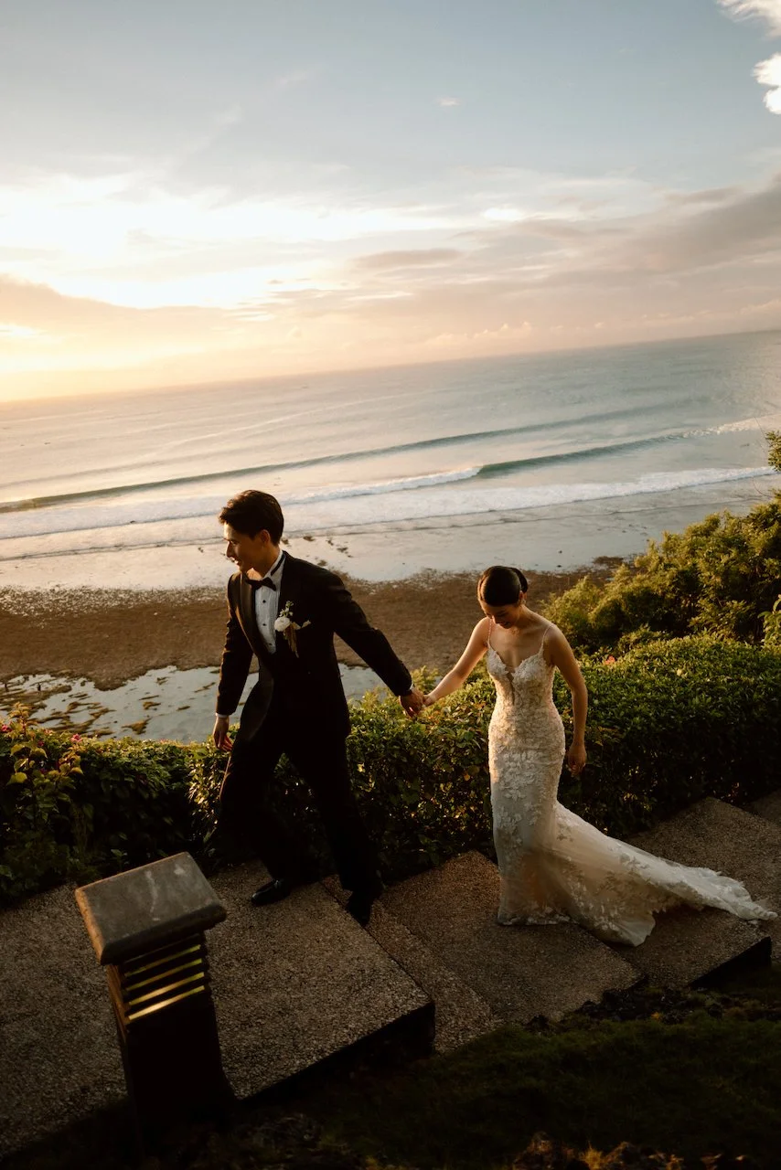Newlyweds holding hands on stone steps overlooking the Bali coastline.