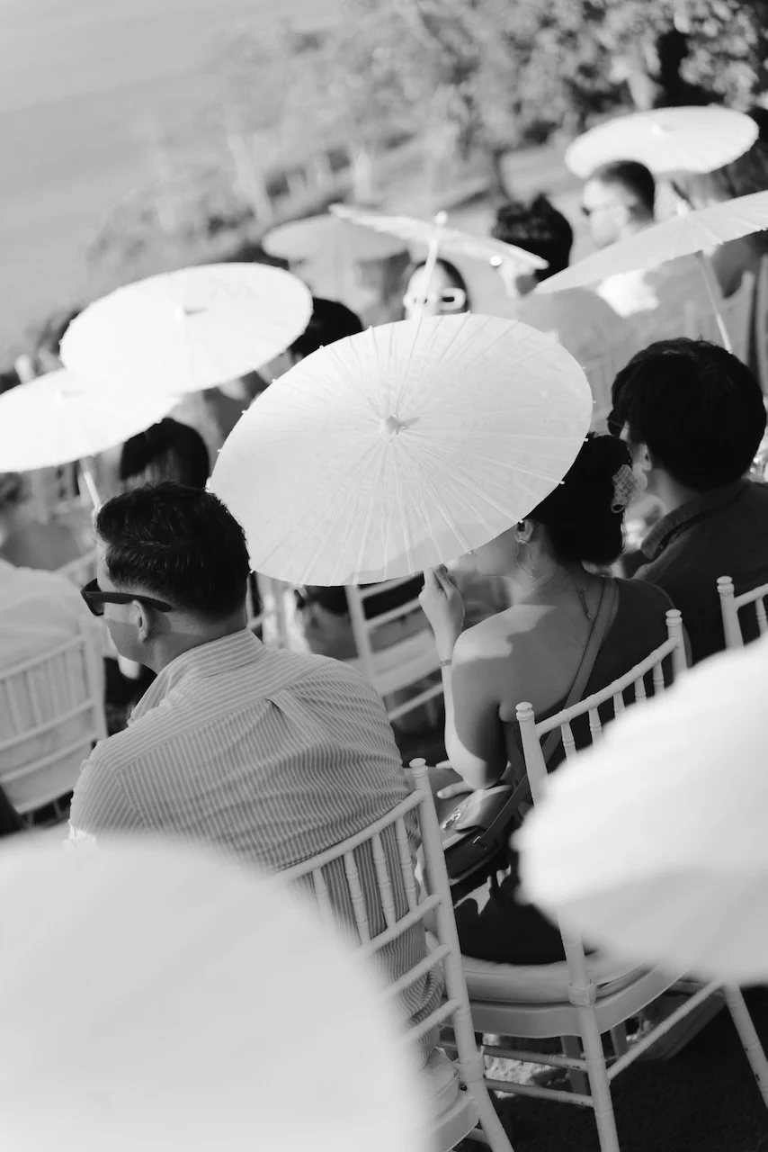 Black and white photo of wedding guests holding white umbrellas for shade during the ceremony