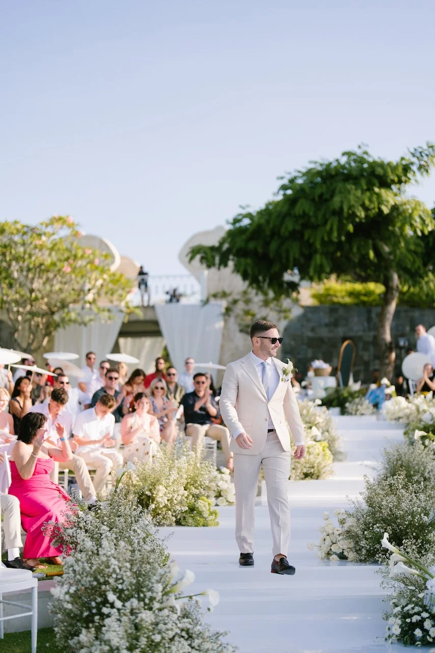 Groom Thomas walking down the white aisle lined with lush white florals