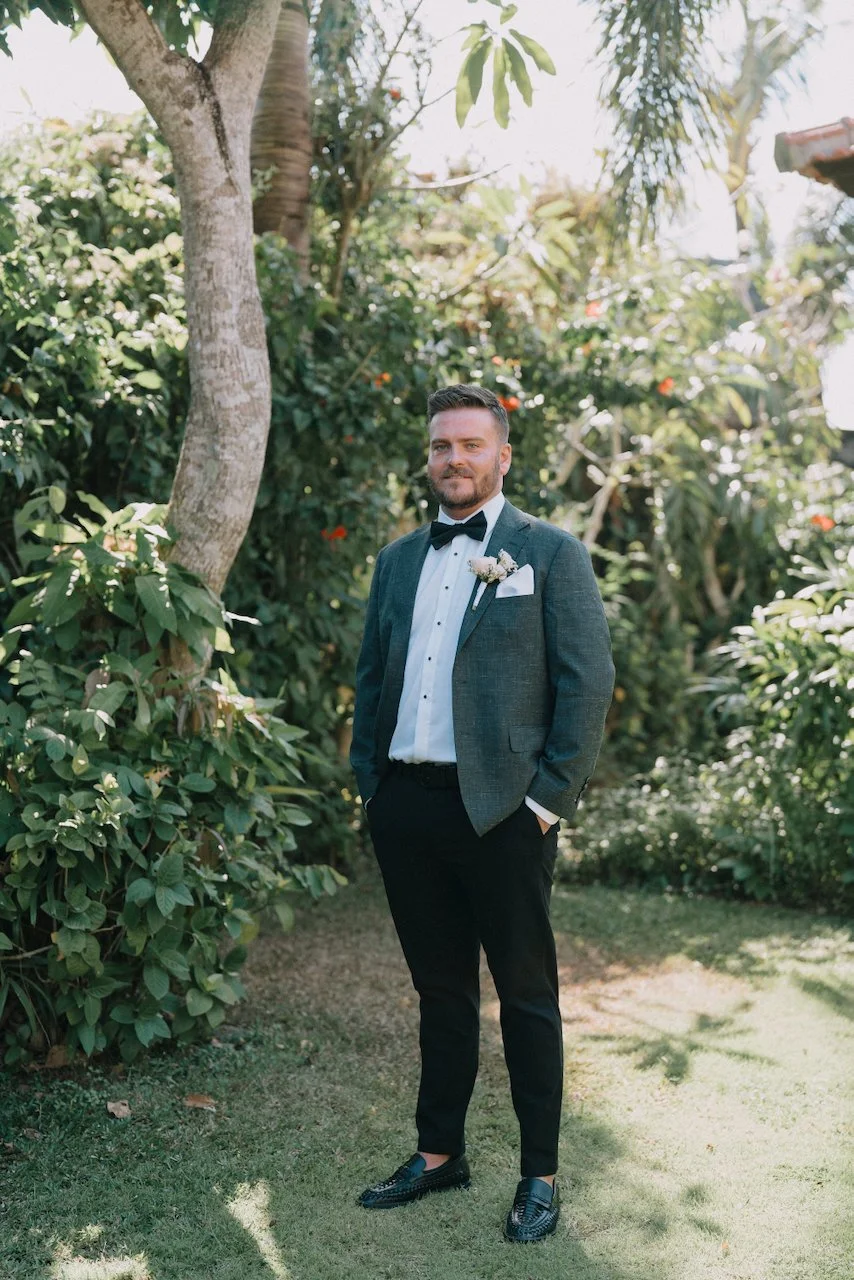 Groom Liam standing in a tropical garden wearing a dark green suit jacket and black bow tie