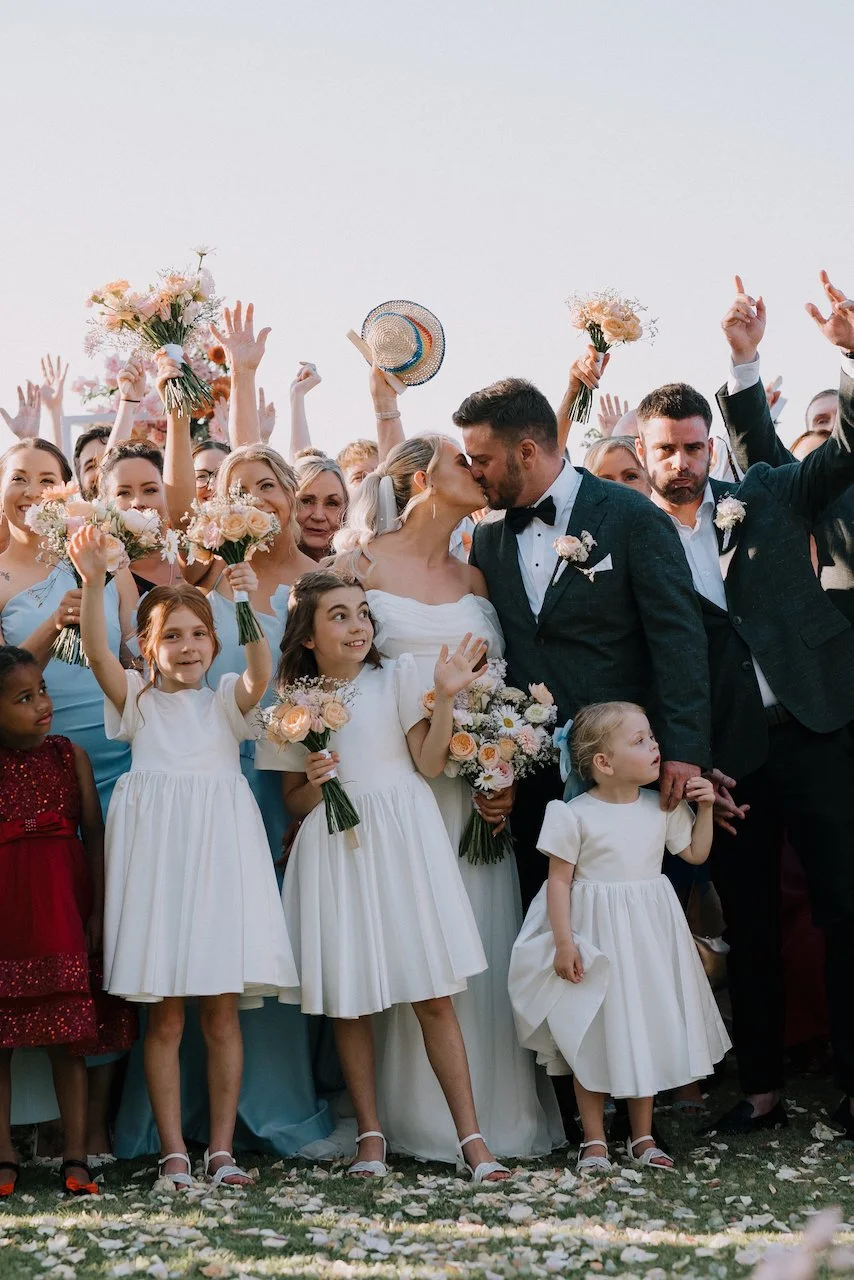 Large group photo of wedding guests cheering with the bride and groom on a clifftop lawn