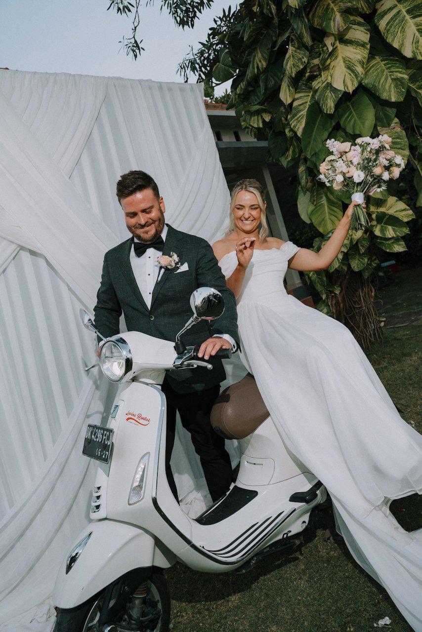Tegan and Liam smiling while sitting on a vintage white Vespa scooter in their wedding attire