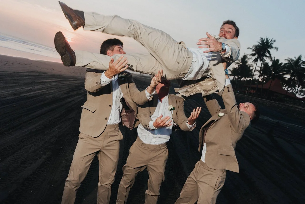 Groomsmen tossing groom Devin into the air on a Bali black sand beach at sunset