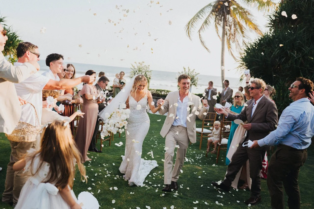 Happy wedding guests throwing white flower petals as the newlyweds walk down the aisle