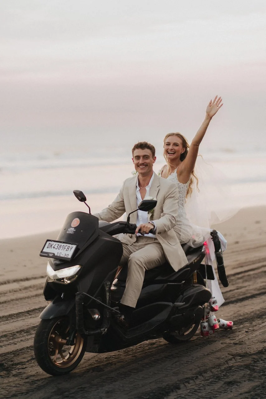 Bride and groom cheering with one hand up while riding a scooter on the beach