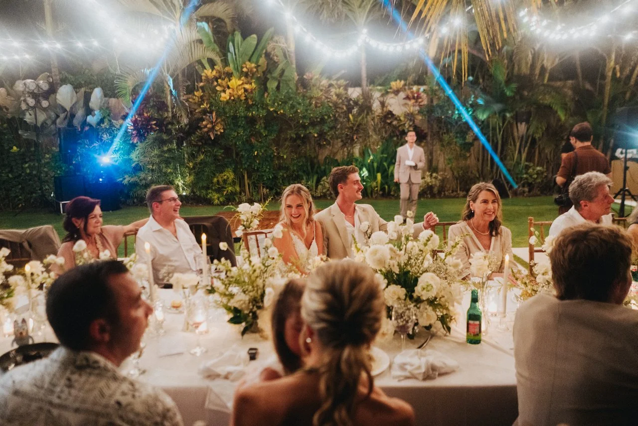 Wedding guests enjoying an open air dinner at a long table decorated with white florals