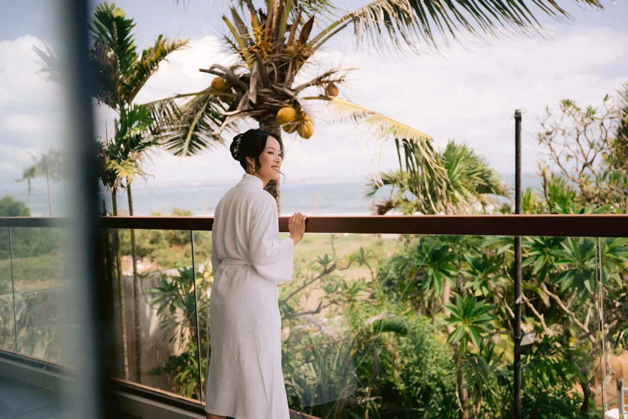 Bride Felicia in a white robe standing on a balcony overlooking the ocean in Bali