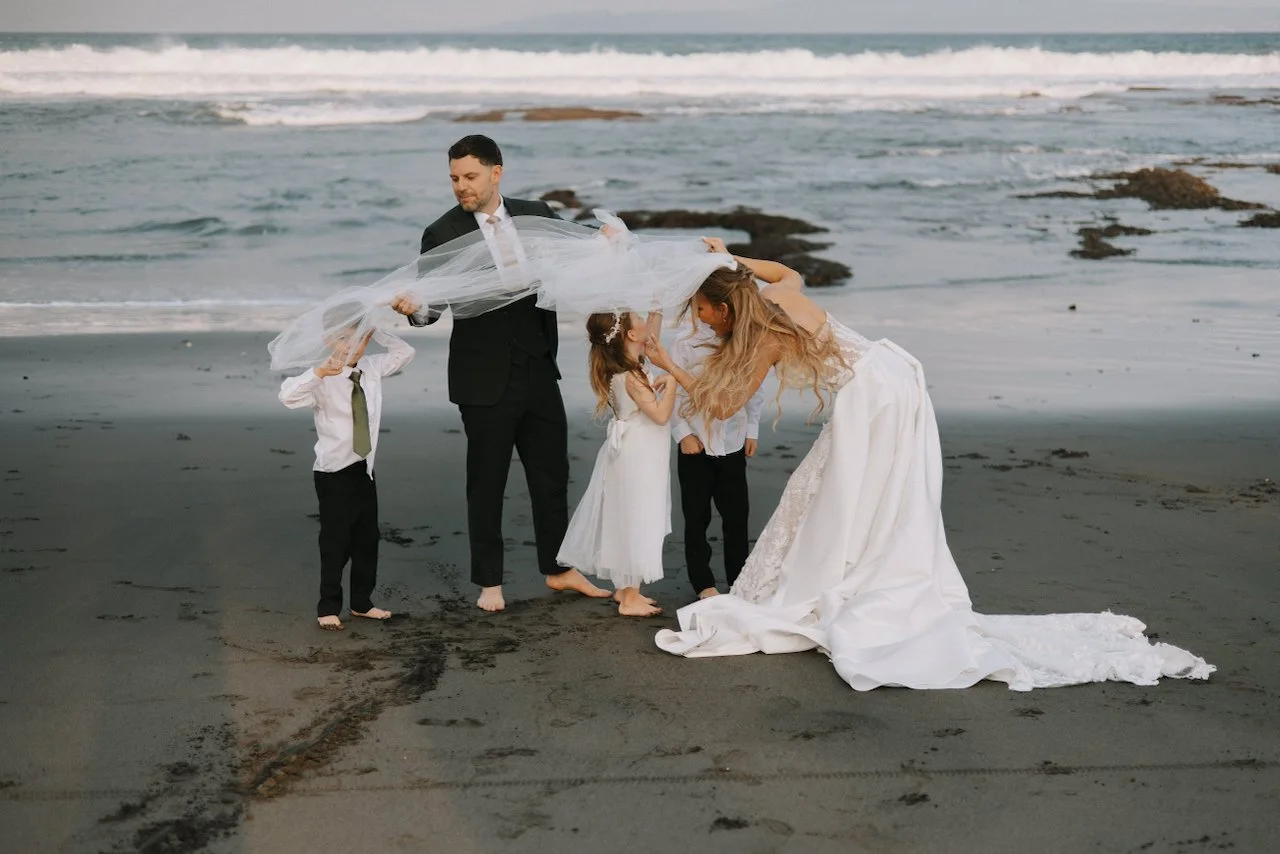 Groom David holding a long white veil over his bride on a black sand beach