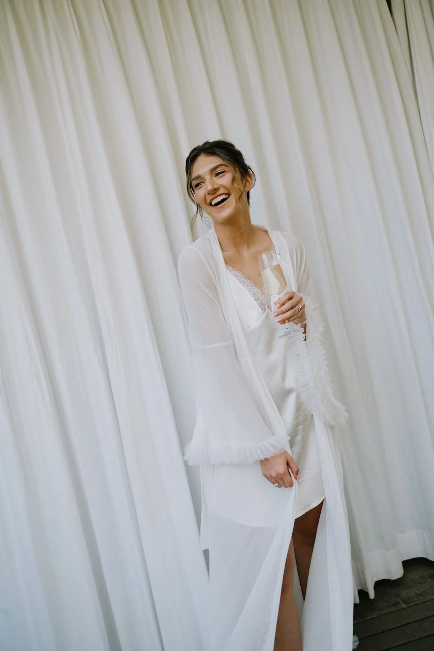 Kelly laughing in a sheer white robe with ruffled sleeves, holding champagne before her Wonderland wedding