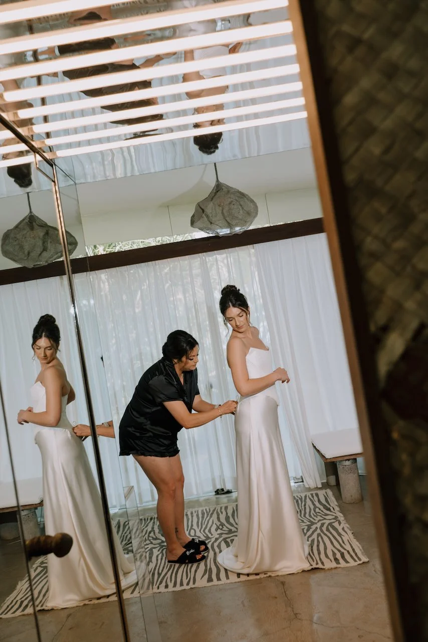 A bridesmaid in a black robe helping Kelly into her satin wedding dress, reflected in a mirror