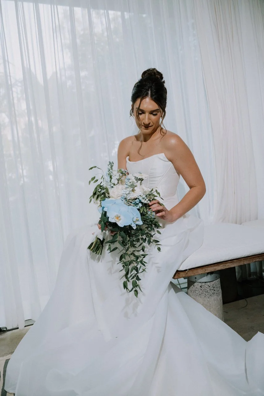 Kelly seated in a corseted white gown holding a pale blue orchid and eucalyptus bouquet