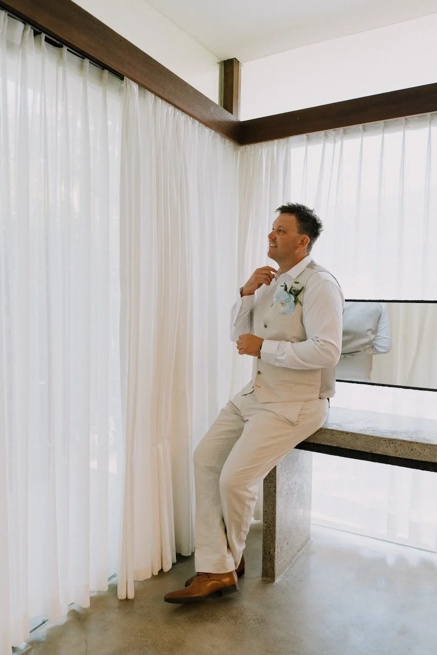 Matt in a linen waistcoat and white shirt with a blue orchid boutonniere, looking out beside white curtains