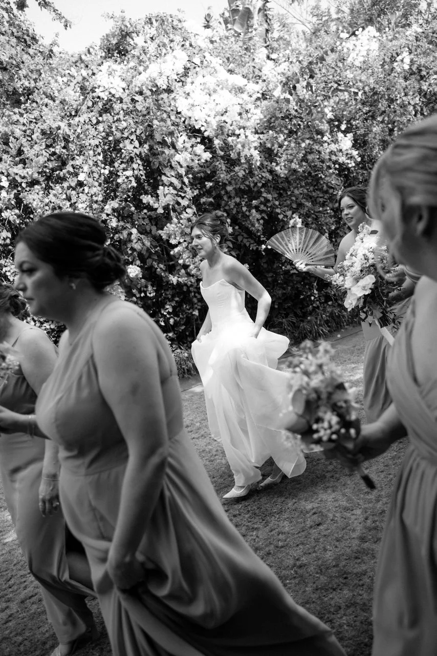 Black and white photo of Kelly lifting her dress as bridesmaids walk past bougainvillea, one holding a fan