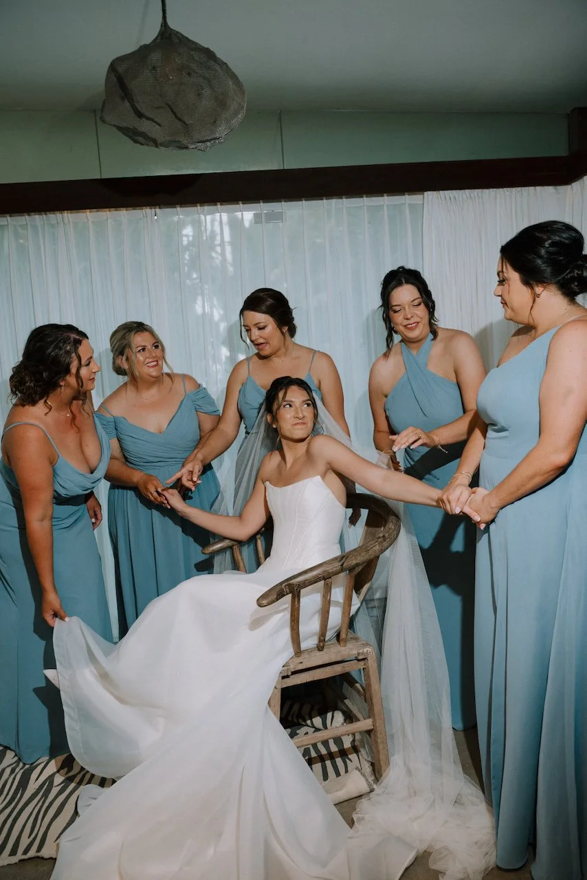 Kelly seated in a wooden chair holding hands with bridesmaids in dusty blue dresses