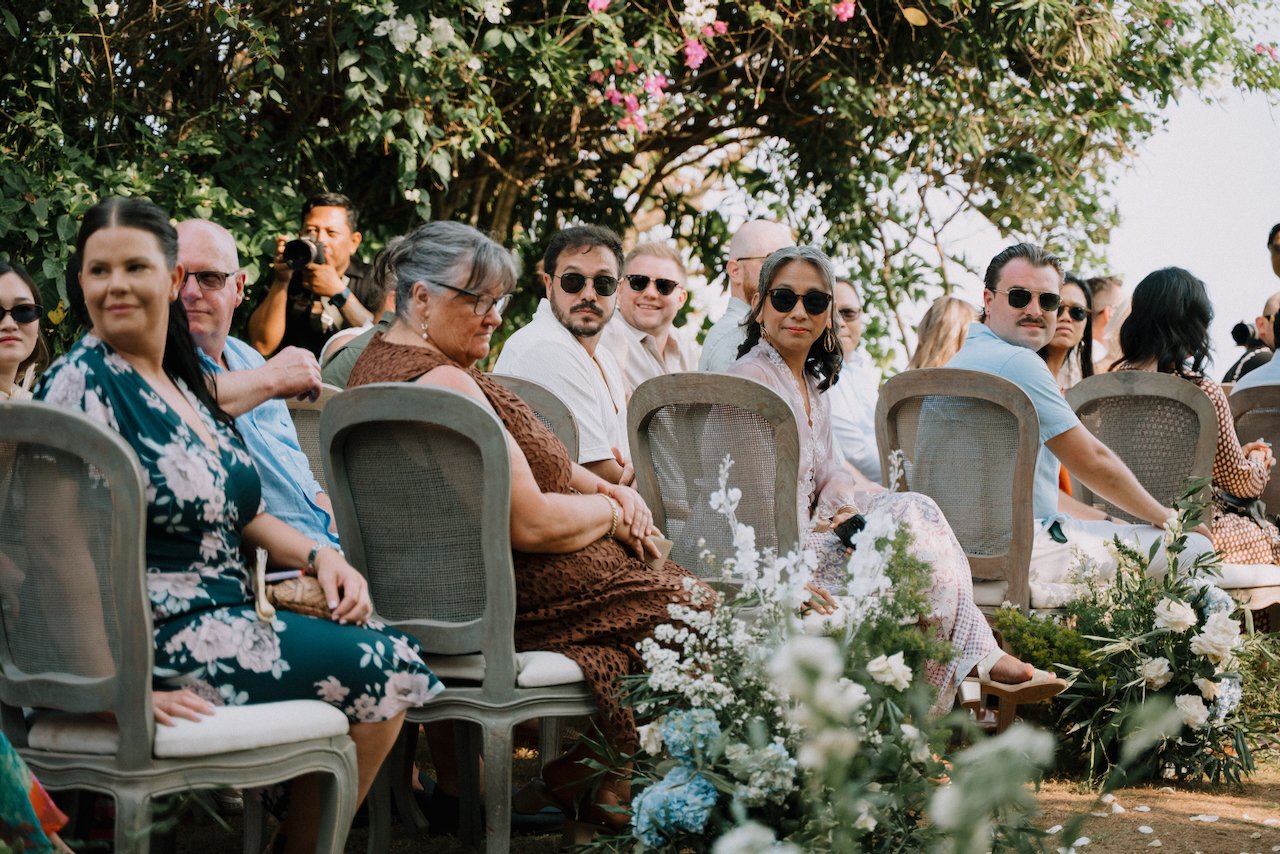 Wedding guests seated on rattan chairs among white and blue flowers, with a photographer behind them