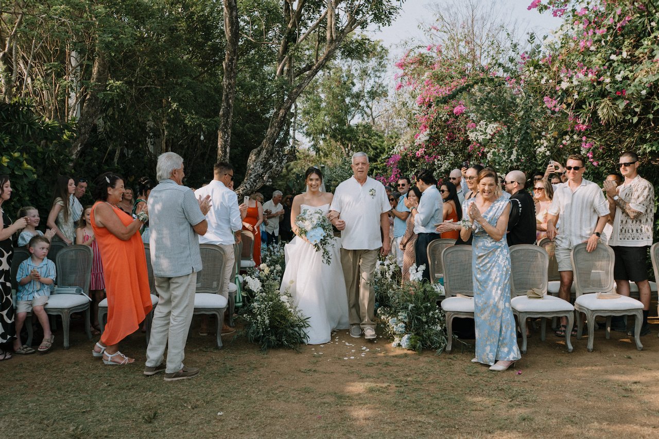 Kelly walking down the aisle with her father as guests applaud under bougainvillea trees